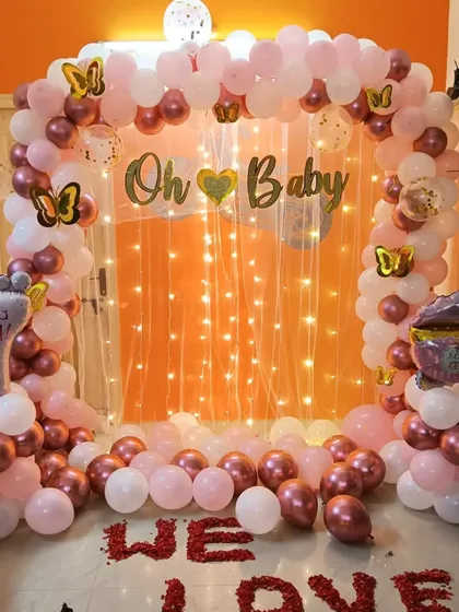 A beautiful and simple baby shower decoration for a home setting. It features a balloon arch in rose gold and pink against a fairy light curtain, with 'Oh Baby' signage and themed foil balloons.