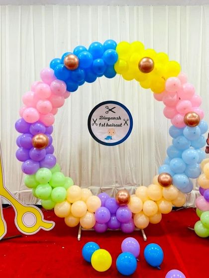 A colorful balloon ring for a Mundan (first haircut) ceremony. The setup includes a large scissor prop and a sign for the baby's milestone.