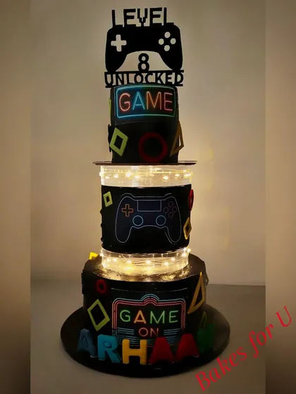 A cool, illuminated gaming-themed cake for an 8th birthday. This multi-tier cake features neon-style gaming signs and controllers, with lights in the middle tier for a glowing effect.