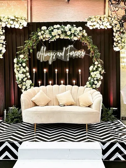 A chic and classic couple seating area for a glam Sangeet. The "Always and Forever" neon sign, circular floral arrangement, and bold black-and-white chevron stage create a modern and stylish focal point.