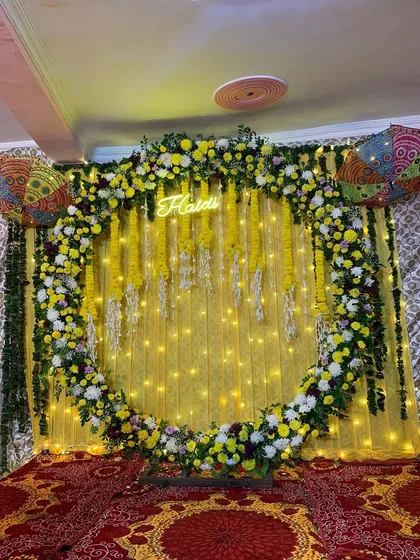 A vibrant and modern Haldi backdrop. This setup features a full circular arch covered in yellow and white flowers, with a custom 'Haldi' neon sign glowing in the center against a bright yellow drape and fairy lights.