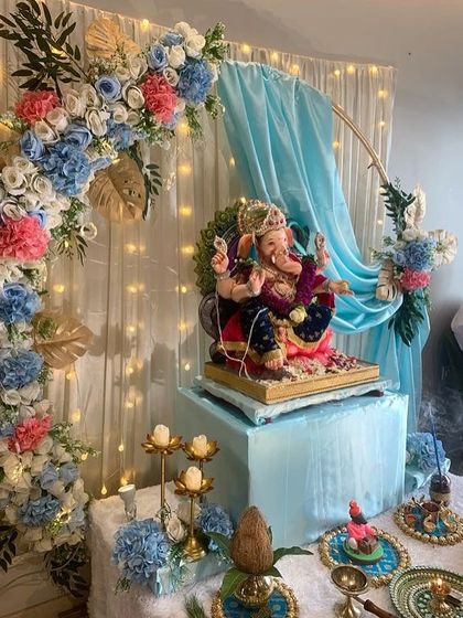 A beautiful blue and pink themed Ganpati decoration. The floral arch is accented with golden leaves, and the light blue fabric draping creates a soft and dreamy background.