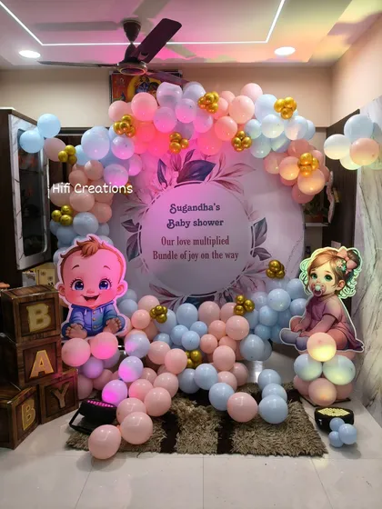 A sweet and simple baby shower decoration for a home party. The setup includes a pastel pink and blue balloon arch, a personalized backdrop, and "BABY" display boxes.