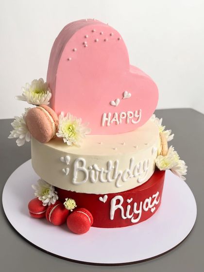 A celebration layered in love. This two-tiered birthday cake features a unique criss-cross style, a heart-shaped top tier, and a beautiful blend of blush pink, ivory, and deep red, finished with fresh flowers and macarons.