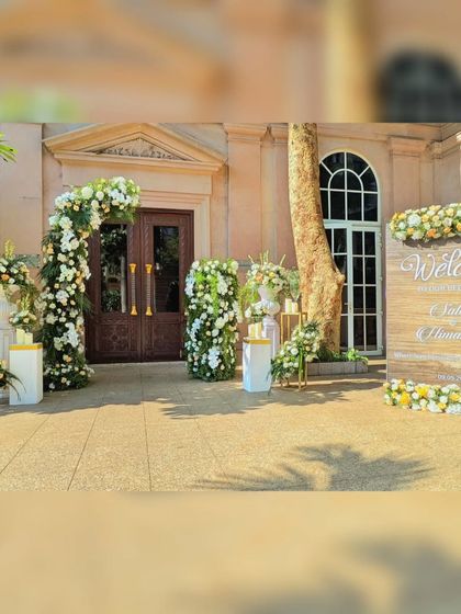 A full view of the outdoor welcome area, showing the floral arch and the rustic wooden welcome sign. This setup creates a charming and inviting atmosphere for a daytime wedding.