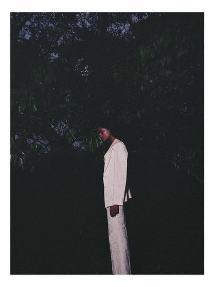 A low-light, moody shot for Sruti Dalmia. The styling of the light-colored suit against the deep darkness of the woods at dusk creates a high-contrast, enigmatic image that focuses on silhouette and atmosphere.
