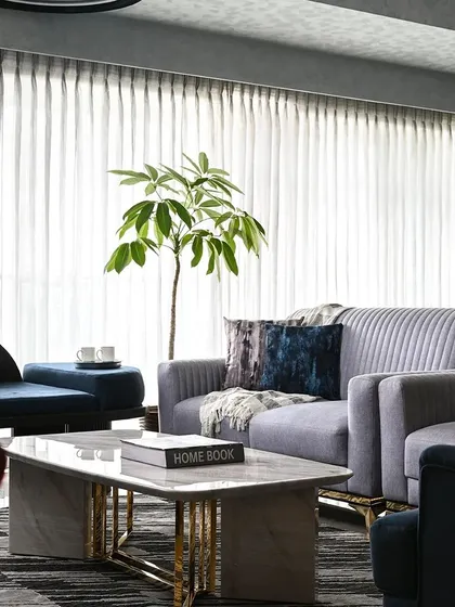 A serene living room where a neutral grey palette is enlivened by deep blue accents. The space is unified by luxurious materials, including a marble coffee table with brass legs and plush, fluted sofas, all set against sheer curtains that bathe the room in soft, natural light.