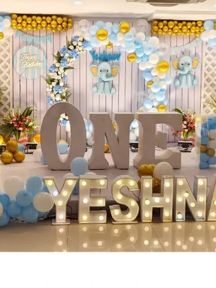 A grand first birthday celebration for Yeshnav. The stage is decorated with a massive balloon backdrop in blue, white, and gold, with large light-up letters spelling out the name for a truly show-stopping effect.