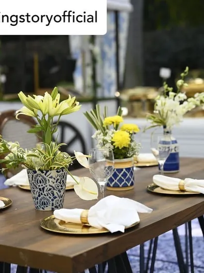 A charming and rustic table setup for an outdoor event. The combination of wooden tables, fresh yellow and white flowers, and gold charger plates creates a warm and inviting atmosphere.