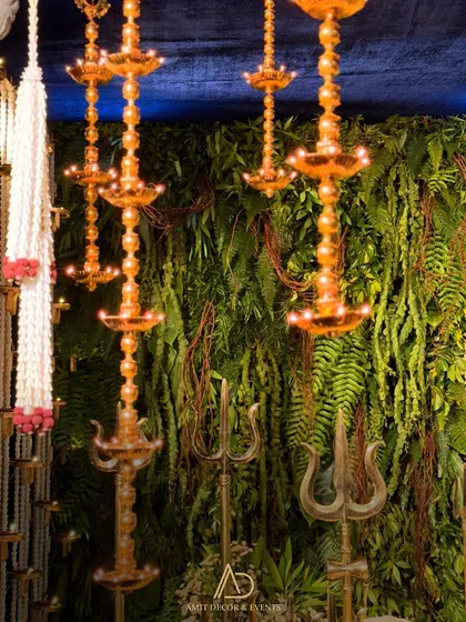 Hanging brass diyas and trishuls set against a lush green wall, creating a beautiful contrast of textures and materials.