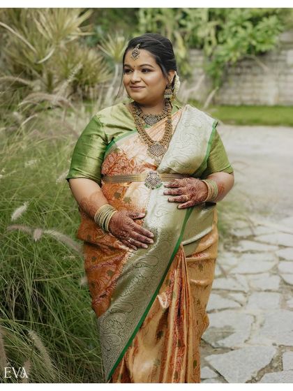 A beautiful three-quarter portrait of an expectant mother in a traditional silk saree, standing in a garden. Her hands rest on her belly, highlighting the intricate jewelry and the glow of pregnancy.