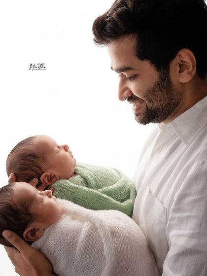 A father smiling down at his two newborn twins, one in a white swaddle and one in green.