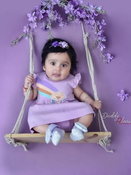 A sweet baby girl in a purple dress sits on a small wooden swing decorated with lavender flowers, all against a matching purple backdrop. This creates a simple yet beautiful color coordinated photo.