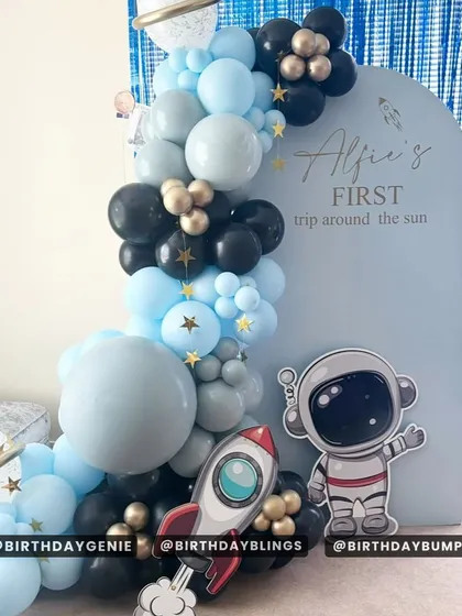 A "First Trip Around the Sun" theme with a clean, minimalist design. The light blue backdrop, simple balloon garland, and cute astronaut and rocket cutouts are perfect for a modern first birthday.