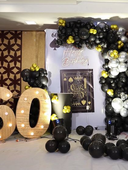 A casino or card-themed 60th birthday decoration. This unique setup uses a black and gold balloon arch, a custom backdrop with a playing card design, and large rustic marquee numbers.