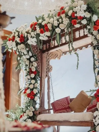 A close-up of the floral arch over the traditional jhoola, featuring a mix of red and white roses with fresh green foliage. The hanging jasmine flower chandelier adds a fragrant and beautiful detail.