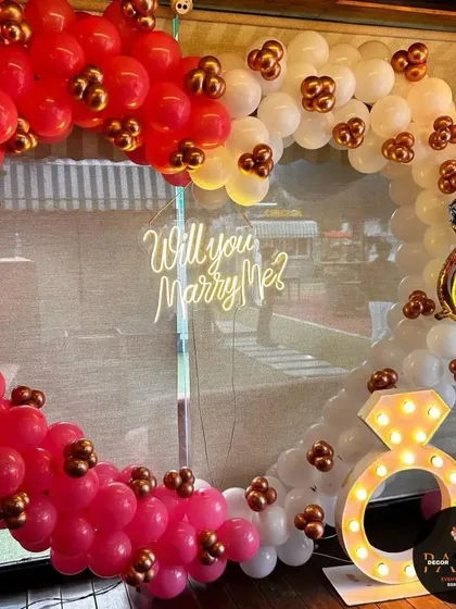 This is a fun and modern proposal setup I created, featuring a heart-shaped balloon arch in red and white. A "Will you Marry Me?" neon sign and a large, light-up diamond ring prop add a playful and trendy touch to the decoration.