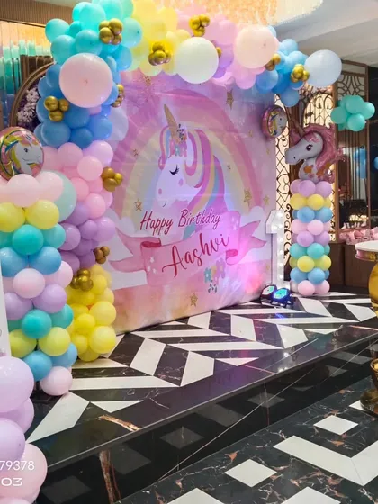 A magical unicorn-themed birthday party setup. This decor includes a pastel rainbow balloon arch over a unicorn backdrop, with a matching unicorn balloon stand.