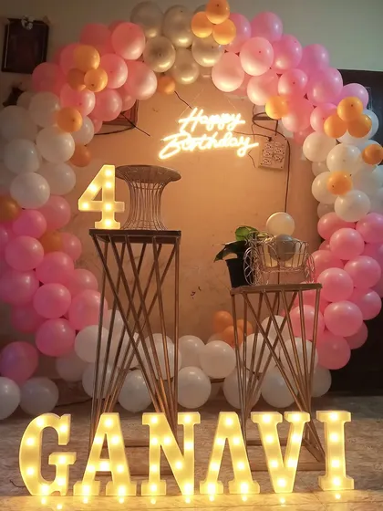 A fourth birthday party decoration in pink and white. This setup includes a circular balloon arch, a neon sign, and large light-up marquee letters for the name.