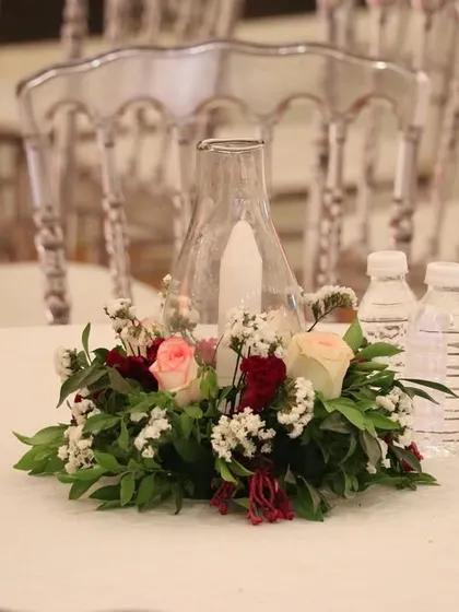 A simple yet beautiful table centerpiece featuring a candle in a glass hurricane lamp, surrounded by a wreath of fresh red and white flowers.