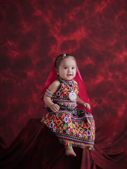 A full-length view of the baby's adorable Navratri outfit. The rich red background makes the vibrant colors of her dress pop, creating a beautiful and festive baby photograph.