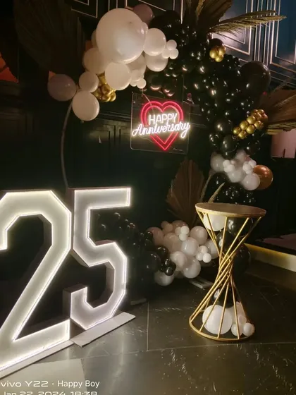 A luxurious 25th anniversary backdrop in a club setting. The decor features a black, white, and gold balloon arch with dried palm leaves, a red 'Happy Anniversary' neon sign, and large '25' marquee numbers.