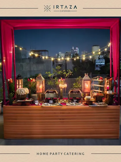 A stunning rooftop home party catering setup. The food station is framed by vibrant pink curtains and string lights, with the city skyline in the background, creating an unforgettable urban party vibe.