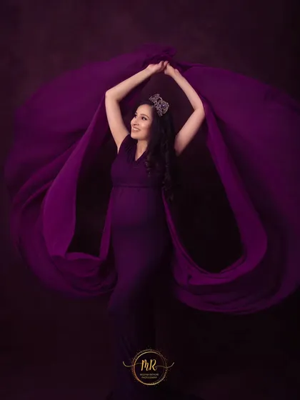 A powerful pose with flowing purple fabric. The mother-to-be wears a crown and holds the fabric aloft, creating a dynamic and regal maternity portrait.