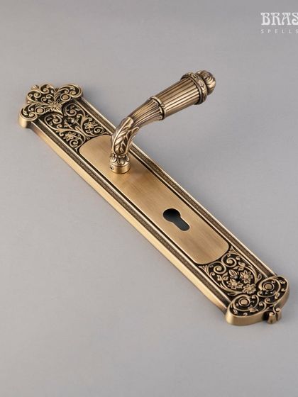 The Monsella door lever in an antique brass finish. The handle's form is complemented by an ornamental backplate with intricate floral and scroll details, making it a statement of artisan craftsmanship.