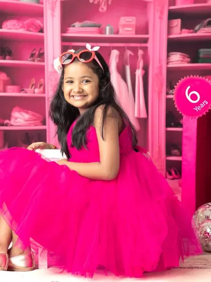 A six-year-old girl smiles brightly in her Barbie-themed shoot, sitting beside a life-sized Barbie box in a room filled with pink.