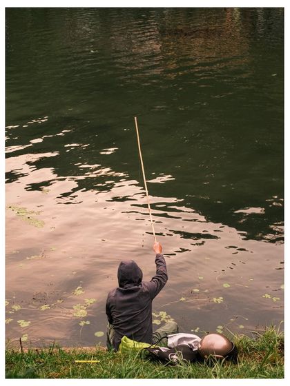 A person seen from behind, fishing with a simple bamboo rod at the edge of a lake. The focus is on the quiet patience of the act.