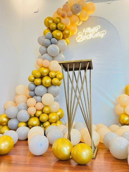 This partial balloon arch in pastel peach, grey, and chrome gold creates a modern and sophisticated look for a birthday party. The setup includes a sleek golden pedestal for the cake.