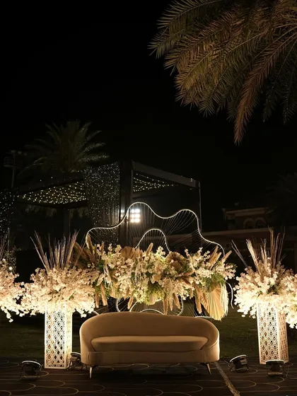 An elegant and modern sangeet stage setup. The design features clean lines, beautiful white floral arrangements, and a backdrop of warm string lights, creating a sophisticated space for performances.