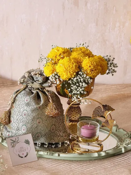 The Golden Aura Tray is a symbol of prosperity and joy. It features a flower vase with marigolds, a candle holder, and a brocade potli filled with our gourmet trail mix.