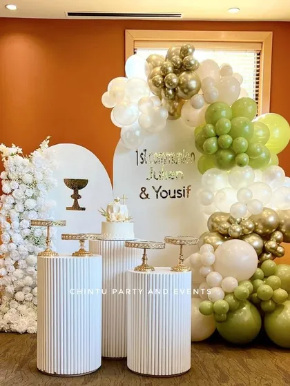 A First Holy Communion backdrop decoration in sage green, white, and gold. The setup includes ridged pedestals and a small white floral tree.