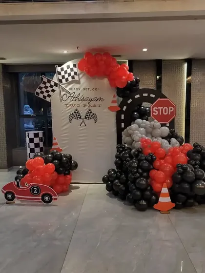 A "Two Fast" car racing theme for a second birthday. The setup includes a custom backdrop with checkered flags, a balloon arrangement in red, black, and grey, and a cutout of a race car.