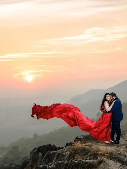 A breathtaking pre-wedding portrait captured during sunset over the hills. The flowing red gown adds a touch of drama against the vast landscape, creating a truly cinematic and romantic photograph.