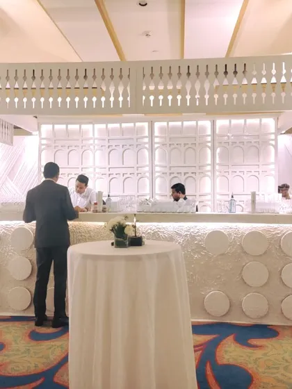 A stunning, custom-designed white bar for a luxury reception. The bar features intricate geometric patterns, a traditional jali-work canopy, and is flanked by large white floral arrangements.
