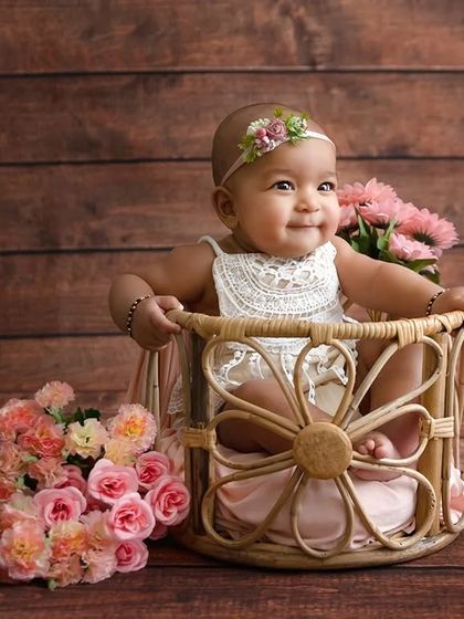 A smiling baby girl sits in a flower-shaped wicker basket, surrounded by pink flowers.