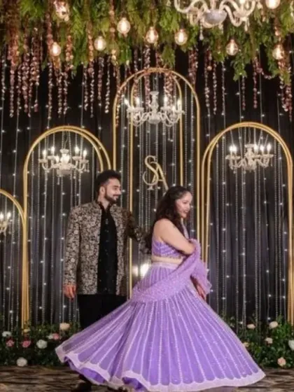 A stunning stage setup for a Sangeet or reception. The backdrop features a curtain of fairy lights, elegant chandeliers, and golden arches, creating a dreamy and glamorous atmosphere.