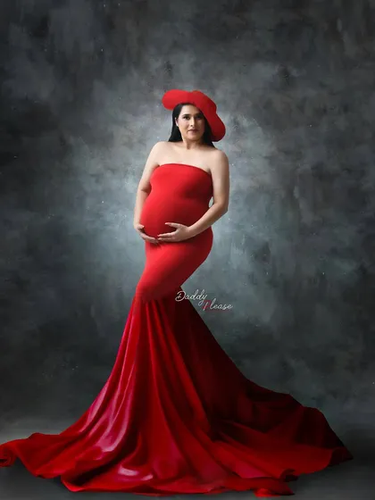 A sophisticated studio shot featuring a red mermaid gown and a matching red hat. The moody, textured grey backdrop adds a touch of classic glamour to this maternity portrait.