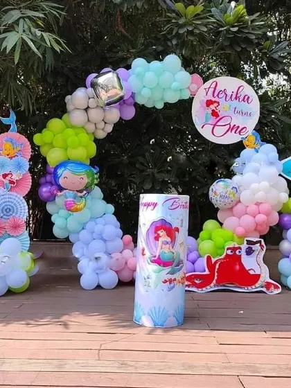 A 'Little Mermaid' first birthday party with an outdoor setup. It includes a colorful balloon arch, a custom-printed plinth, and character cutouts of Ariel and Flounder.