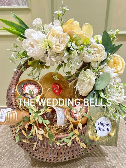 A beautiful and rustic Diwali hamper in a wicker basket. It includes a bottle of wine and other goodies, adorned with a white and yellow floral arrangement.