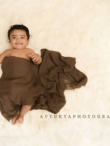 A smiling 3-month-old baby wrapped in a brown cloth on a fluffy white rug.