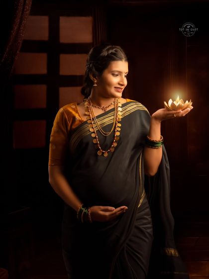 A classic portrait of a mom-to-be in a dark, elegant saree, holding a lit diya that casts a warm glow on her face.
