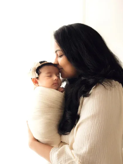 A mother holds her swaddled newborn, kissing her forehead against a brightly lit background. This mother and baby photoshoot captures a moment of pure tenderness.