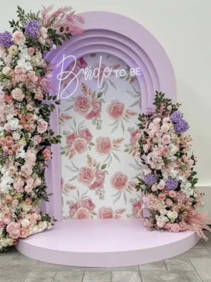 This is a custom-designed "Bride to be" photo booth, perfect for bridal showers or pre-wedding events. The setup includes a lilac-colored arched backdrop with a floral-print interior, a neon sign, and asymmetrical pastel floral arrangements.