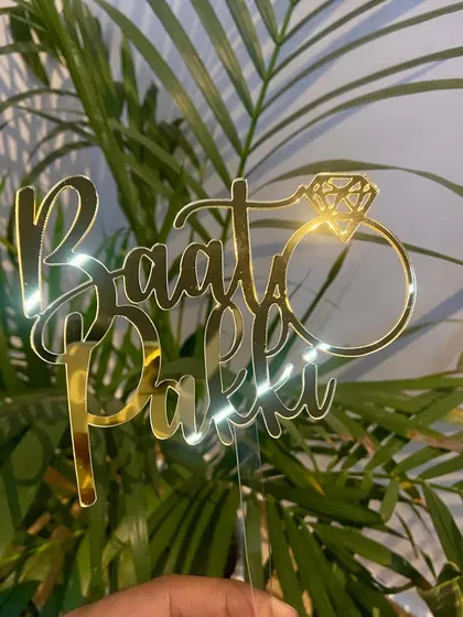 The "Baat Pakki" topper held against a plant, with the light catching the gold finish beautifully.