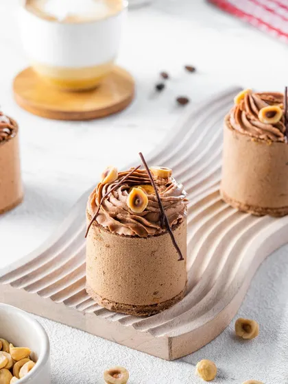 Individual hazelnut mousse desserts on a modern, wavy platter. The bright, airy photograph emphasizes the delicate piping work and textures, presenting them as miniature works of art.