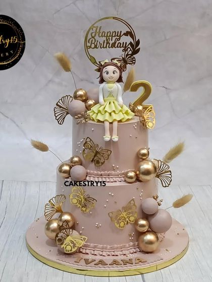 A sophisticated two-tier chocolate cake for a stylish second birthday. It's topped with a cute girl figurine and decorated with golden butterflies, delicate ferns, and shimmering faux pearls for an elegant touch.